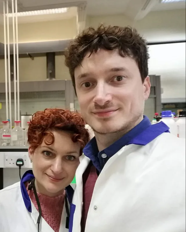 Guy Entract with a fellow chemist in a chemistry laboratory, fume hoods and reagent bottles behind them